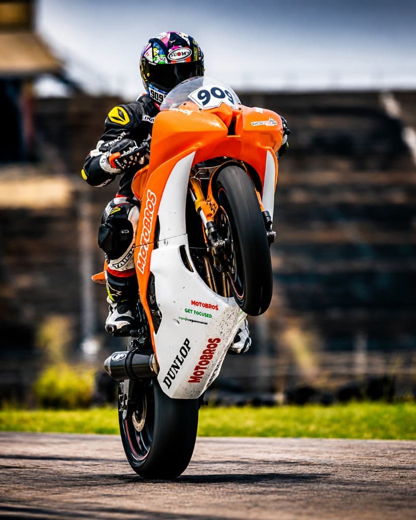 MotoBros rider on an orange and white #909 sportbike performing a vertical wheelie on track